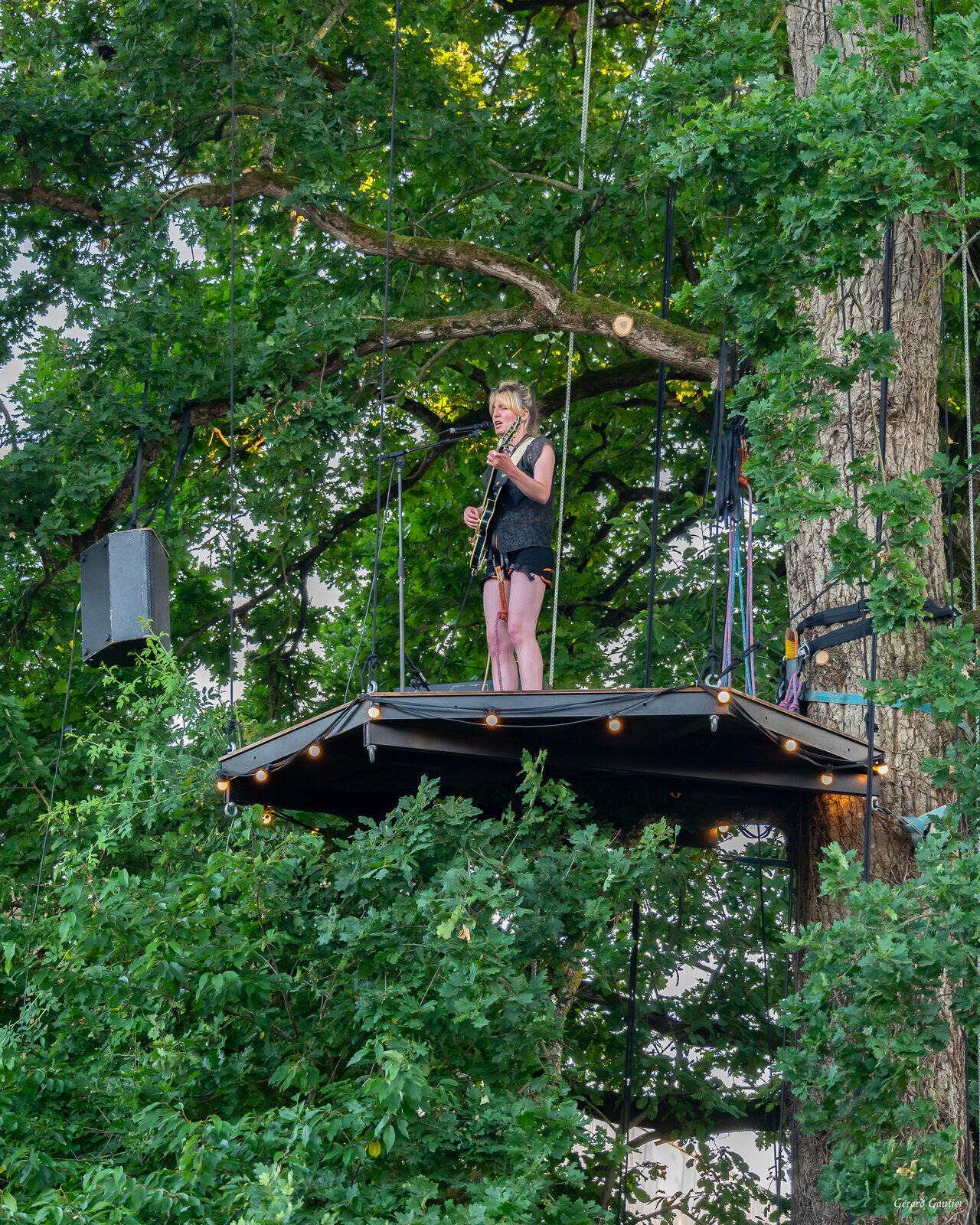 Concert perché sur L'Arborium : chanteuse guitariste sur la scène suspendue dans un chêne avec guirlandes lumineuses - location scène perchée Bretagne